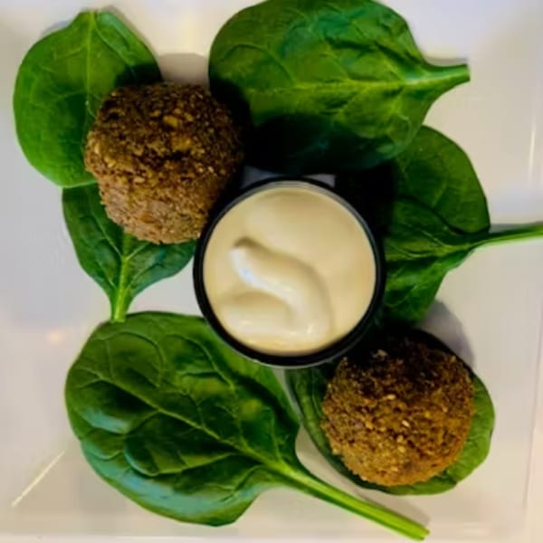 Two falafel balls, a cup of white sauce, and fresh spinach leaves on a white plate.