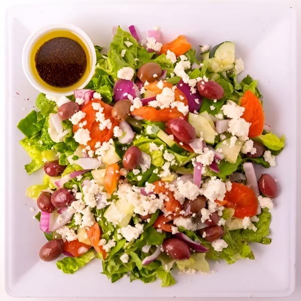 Bowl of Greens' Greek salad with lettuce, tomatoes, olives, feta, and a side of dressing.